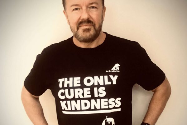 Comedian and activist Ricky Gervais stand wearing a black t-shirt with the caption 'The Only Cure is Kindness'