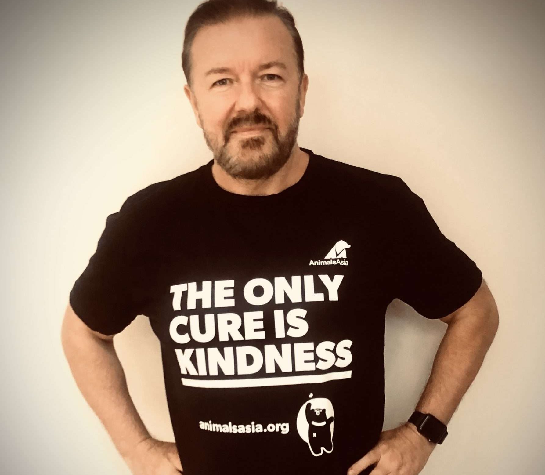 Comedian and activist Ricky Gervais stand wearing a black t-shirt with the caption 'The Only Cure is Kindness'