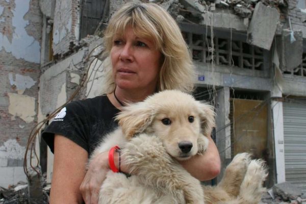 Image of Jill Robinson looking determined and defiant, holding a rescued golden coloured dog from an earthquake zone, with semi-collapsed buildings behind her